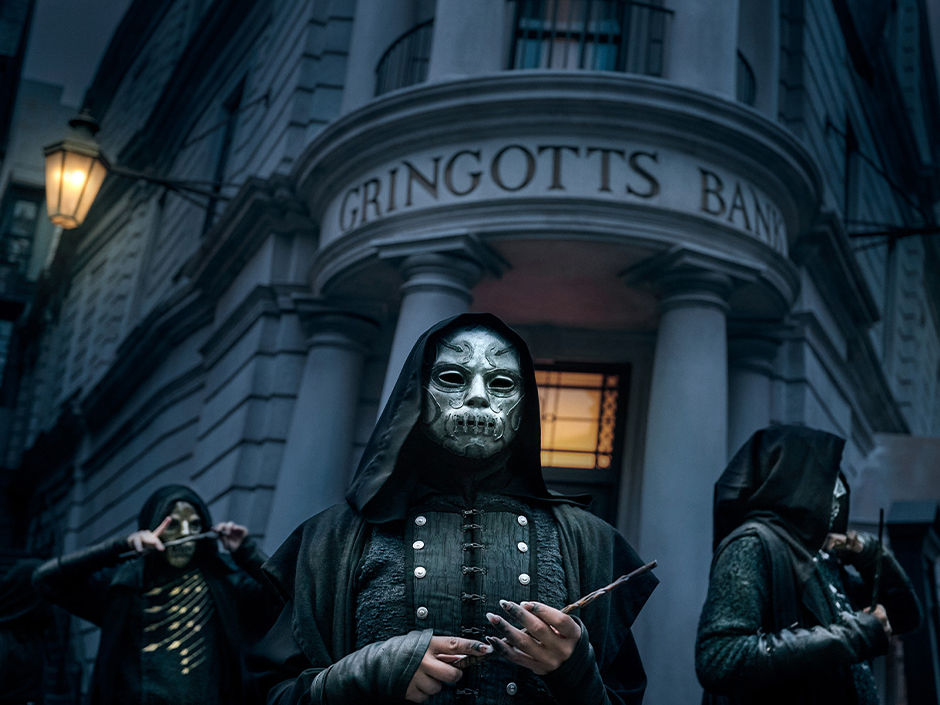 Hooded masked figures outside Gringotts Bank at night, one holding a wand, with an ominous blue-gray mood.