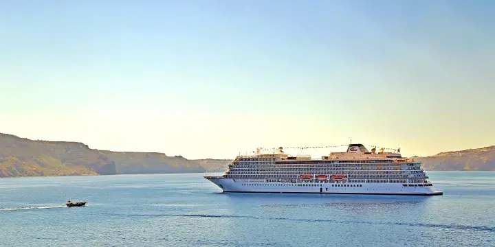 Cruise ship sails in calm blue waters near rocky coast, with a small boat nearby. Clear sky above, conveying a peaceful mood.