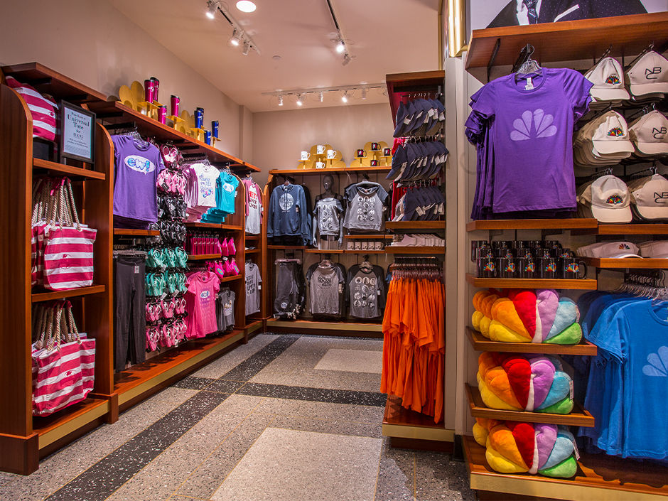 Gift shop with colorful shirts, hats, mugs, and bags. Bright lighting, wooden shelves, red and white striped bags. NBC logo visible.
