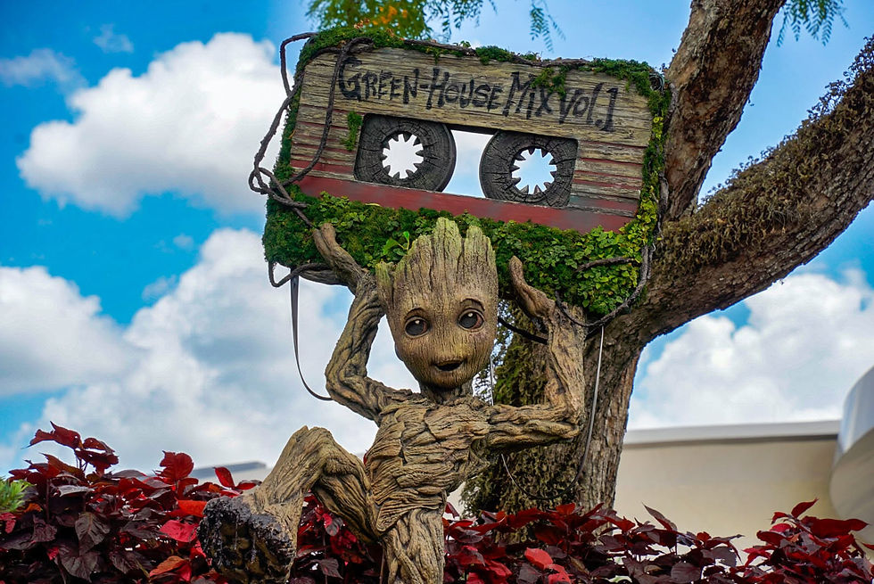 Groot statue sits in a tree holding a cassette labeled GREEN-HOUSE MIX Vol 1, against a blue sky with clouds.