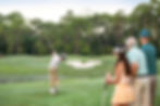 Golfer mid-swing on a green fairway near a white sand bunker, watched by three people, with trees in the background.