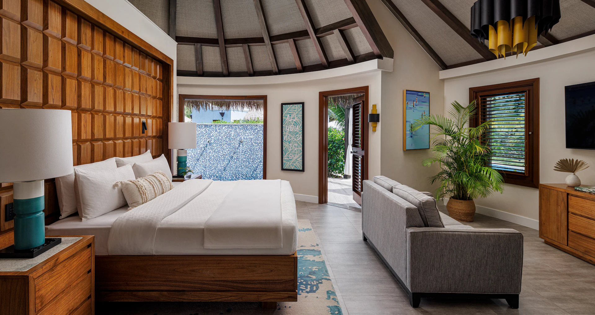 A bedroom with neutral decorations and teal accents. A clean made bed, with all white bedding. A couch sits near the foot of the bed. The window has a tropical view.
