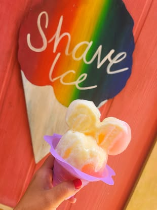 A shave ice shaped like Mickey Mouse in front of a wooden shave ice sign.
