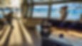 Two people on a ship's bridge; one uses binoculars, the other adjusts a camera. Sunlit room with maps, overlooking glaciers.