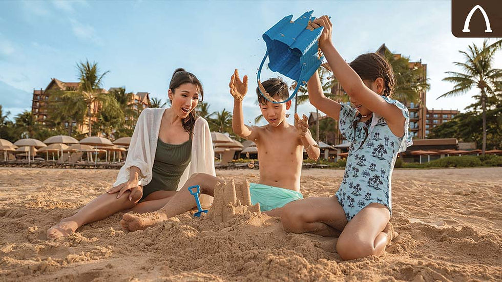 Three children build a sandcastle on a sunny beach. One pours sand from a blue bucket. Palm trees and buildings in the background.