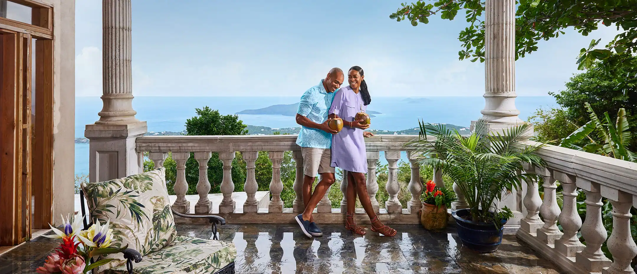 A couple are on a balcony with the ocean in the background, surrounded by tropical plants.