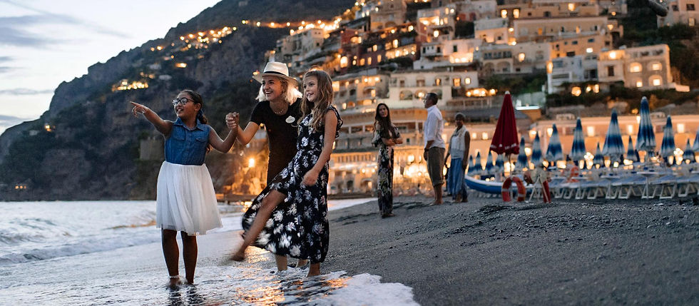 Smiling girls wade at dusk on a beach, with lit hillside houses and blue umbrellas behind them.