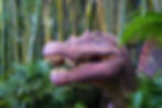 A large dinosaur sculpture with open jaws and sharp teeth surrounded by lush green plants and bamboo in a jungle-like setting.