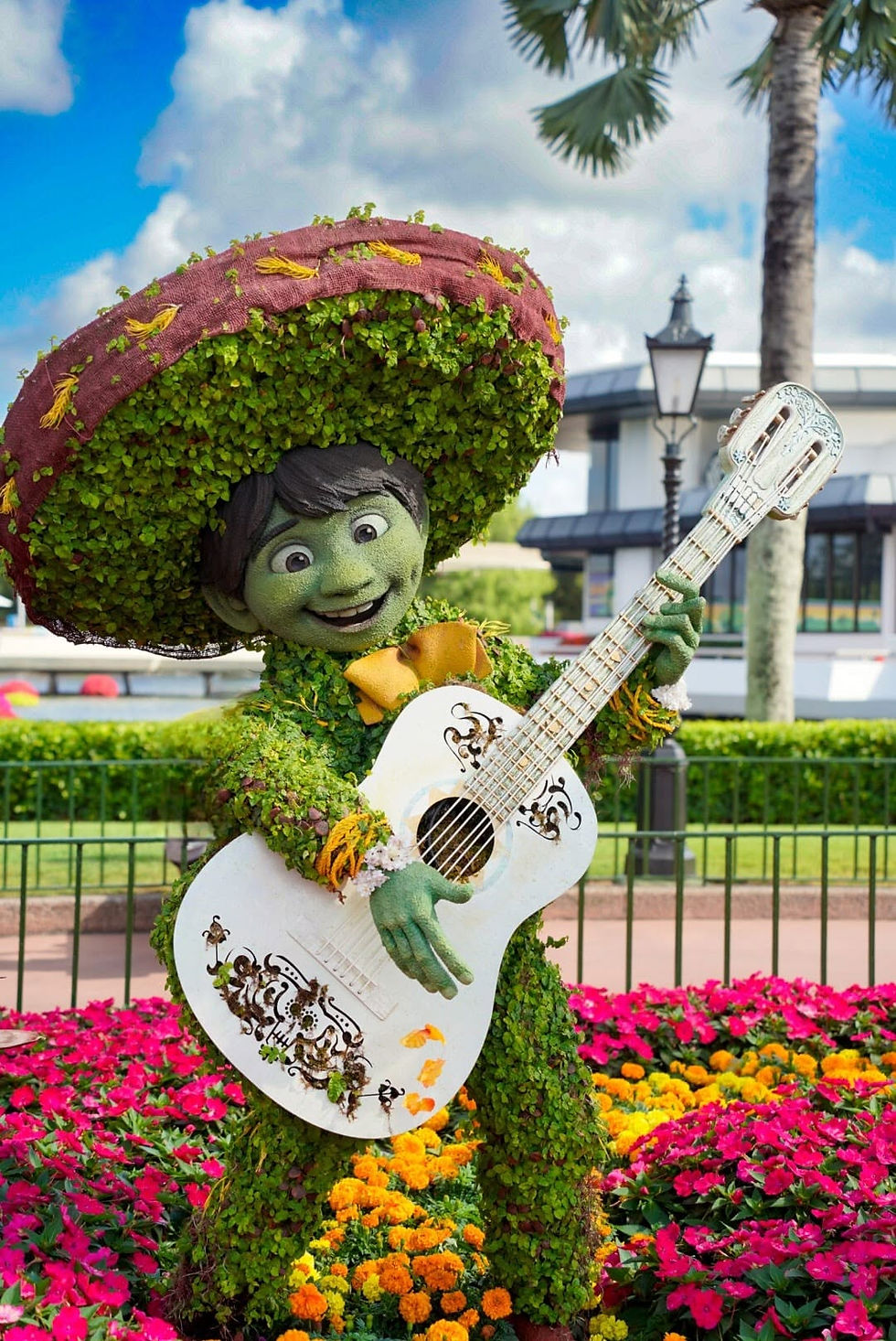 Green topiary child in a giant sombrero plays a white guitar in a bright flower garden under a blue sky