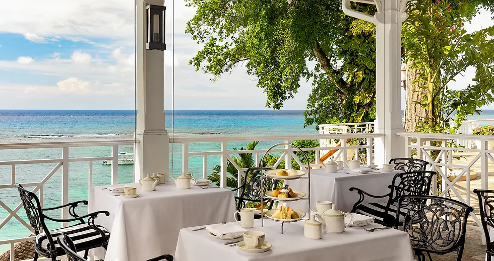 Oceanfront patio with white tablecloths, black chairs, and a pastry stand. Lush greenery and a vibrant blue sea view create a serene setting.