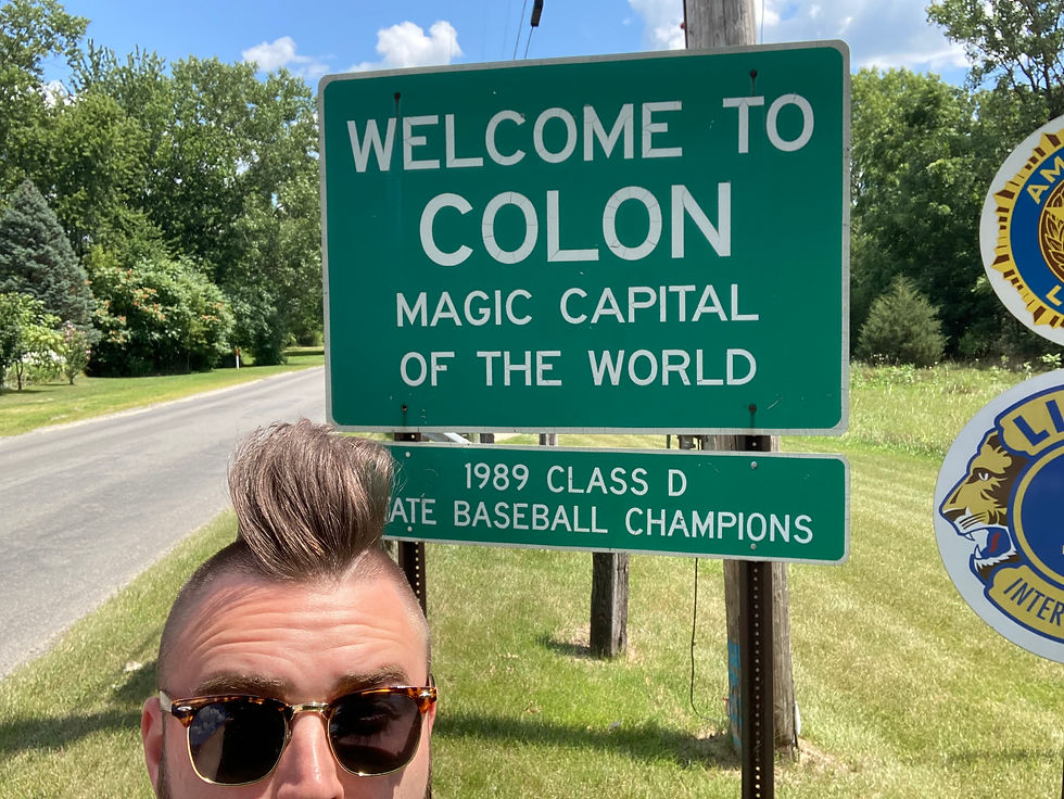 Man with sunglasses and unique hairstyle poses in front of a green road sign reading "Welcome to Colon, Magic Capital of the World," under clear skies.
