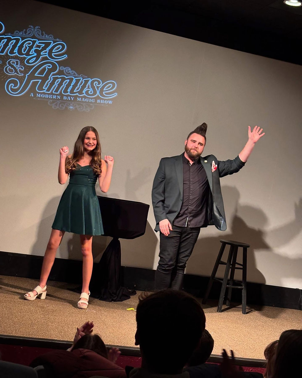 Two people on stage waving to an applauding audience. "Amaze & Amuse: A Modern Day Magic Show" logo in the background.