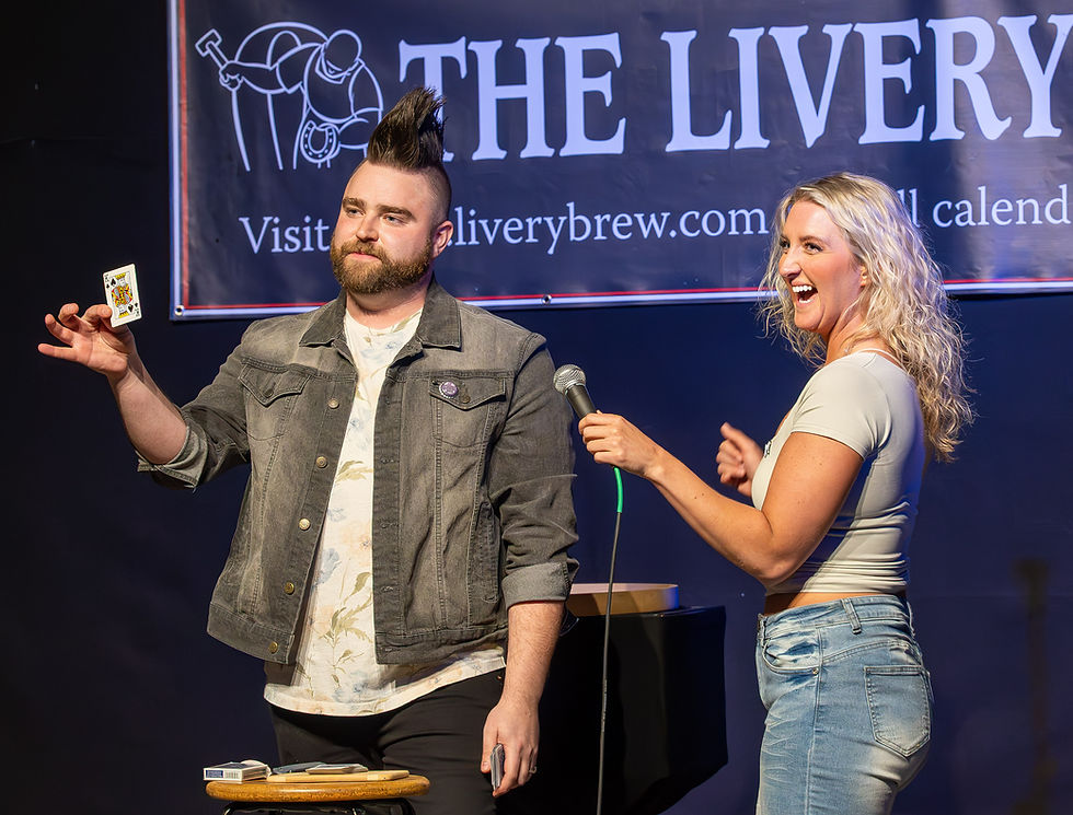 Man with a mohawk holds a card on stage, woman smiles holding a microphone. "The Livery" sign in the background, casual setting.