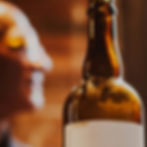 Close-up of a chilled amber beer bottle with a partly visible label, warm bar lights, and a blurred person in the background.