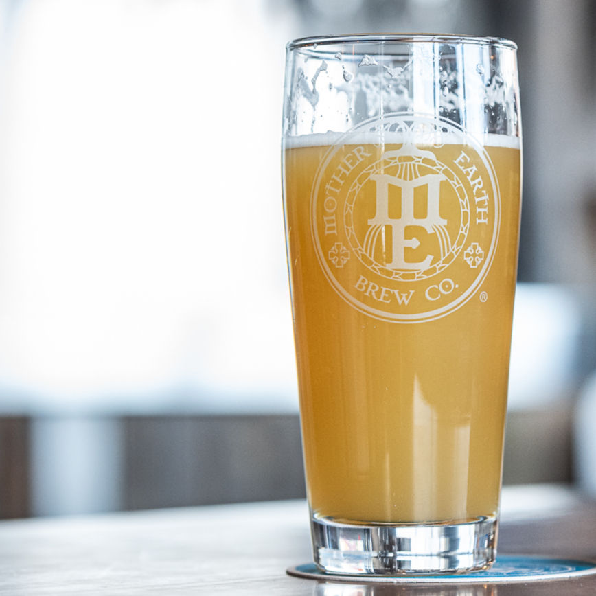 A glass of pale beer with the "Mother Earth Brew Co." logo sits on a wooden surface. Blurred bright background.