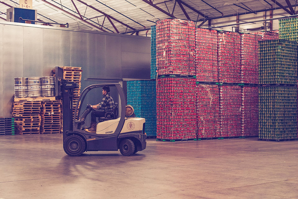 A forklift moves kegs through a brewery with cans in the background.