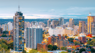 city of Hamilton: City skyline at dusk with illuminated high-rise buildings, greenery in foreground, and a cloudy sky above, creating a serene urban scene.