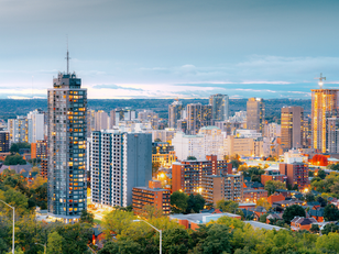 city of Hamilton: City skyline at dusk with illuminated high-rise buildings, greenery in foreground, and a cloudy sky above, creating a serene urban scene.