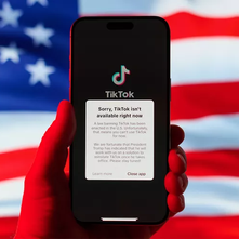 Hand holds a phone displaying TikTok ban notification. Background features a blurred U.S. flag. Red, white, and blue dominate.