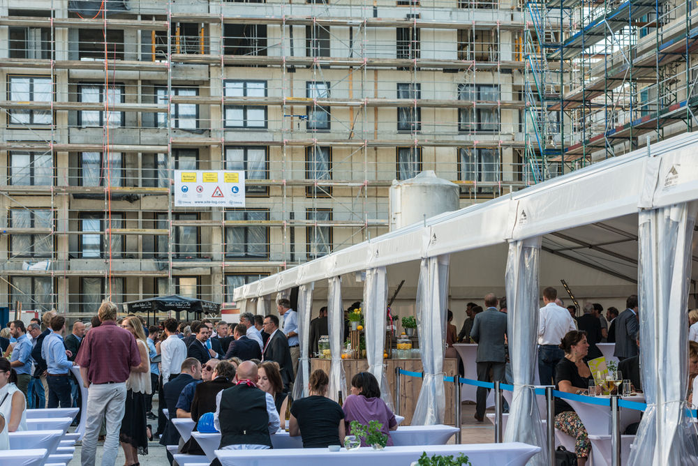 Zahlreiche Gäste feiern unter einem weißen Festzelt direkt neben einem eingerüsteten mehrstöckigen Rohbau ein Baustellenfest.