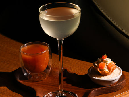 A tall coupe glass of clear cocktail alongside a vibrant michelada chaser and a smoked salmon tartlet, served together on a dark wooden tray at Muro.