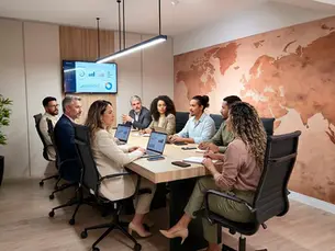 Reunião de equipe em sala moderna, com sete pessoas à mesa, laptops e gráficos na tela, diante de um mapa-múndi na parede.