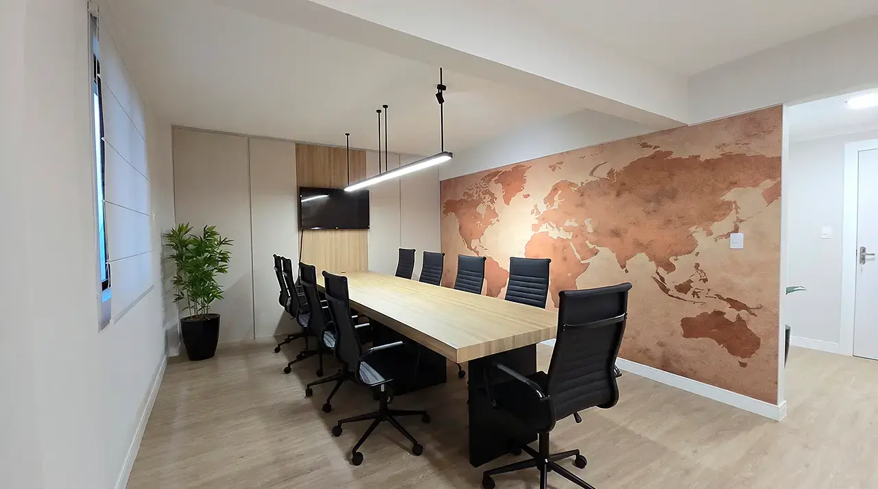 sala de reunião mostrando uma mesa grande com cadeiras e ao fundo uma televisão e um painel. Decoração de planta, luminárias e um papel de parede com o mapa geográfico