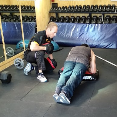 Trainer kneels beside a man holding a plank on a mat in a gym with dumbbells and a mirror, focused training scene.