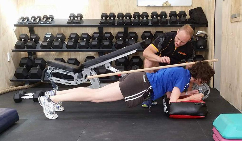 Trainer checks woman holding a plank with a wooden dowel on her back in a dumbbell-filled gym.