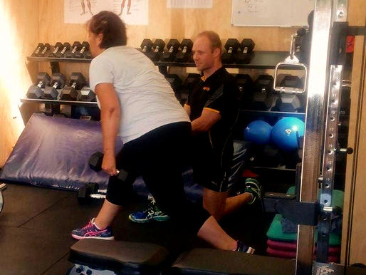 Woman lunges with dumbbells as a trainer kneels beside her in a gym with racks of weights, exercise balls and anatomy posters.