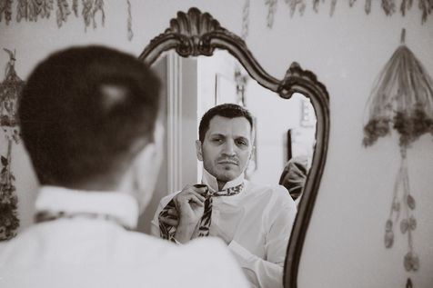 A man adjusts his tie in a mirror as he prepares for his wedding - Julia Bailey