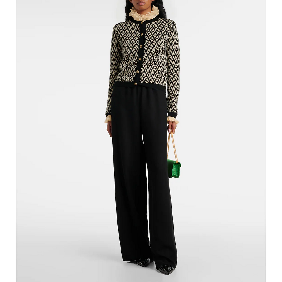 Woman in patterned cardigan and black pants holds a small green purse. The background is plain white, highlighting her outfit.