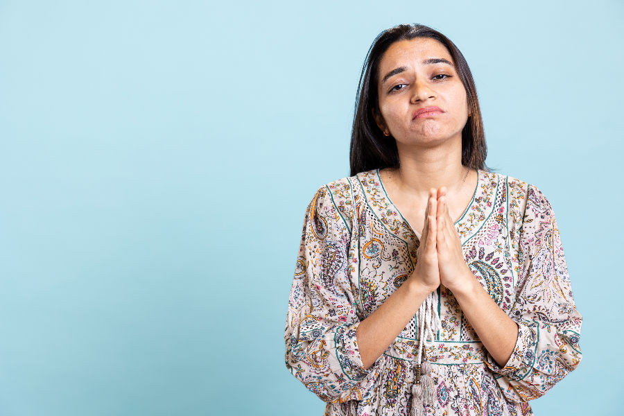 Woman with long hair, wearing a patterned dress, clasps hands in a pleading gesture against a light blue background, looking hopeful.