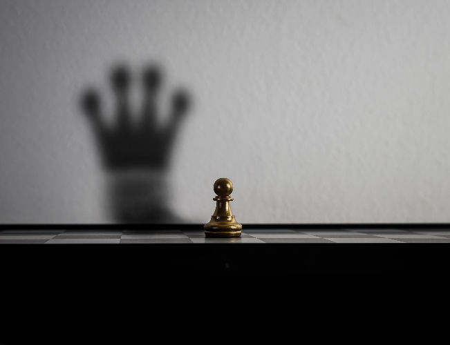 Gold chess pawn on a board with a blurred crown shadow on the wall, creating a dramatic, symbolic mood.
