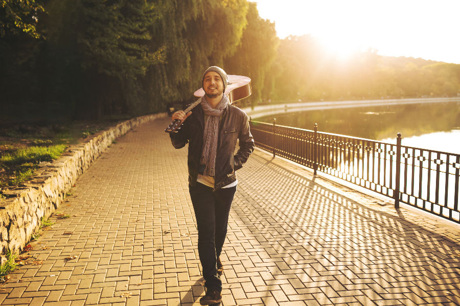 Man walks along a sunlit lakeside path at sunset, carrying a guitar on his shoulder, with trees and water behind.