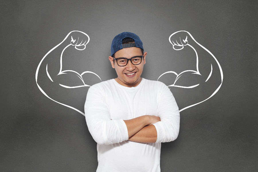 Smiling man in white shirt and cap stands with arms crossed. Chalk muscle drawings on gray background suggest strength and confidence.