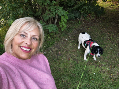 A woman with short blonde hair in a pink sweater takes a selfie outdoors with a black and white dog on a green leash and red harness. The background features green grass and dense foliage.