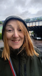 A woman in a hooded jacket stands outdoors on a rainy day near an overpass with blue railings, a wet road, and parked cars in the background.