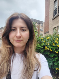 A woman with long hair stands outdoors, in front of buildings and a bush with yellow flowers under an overcast sky.
