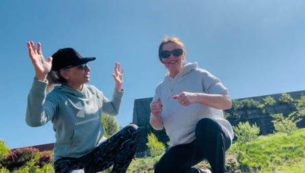 Two individuals outdoors wearing hoodies, one in a light blue Under Armour hoodie with raised hands, the other in a light grey hoodie squatting. The background features greenery and a building under a clear blue sky.