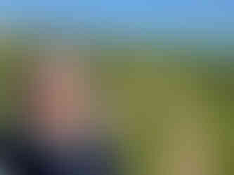 A woman with curly gray hair takes a selfie outdoors on a grassy path leading to a body of water, with clear blue skies in the background.