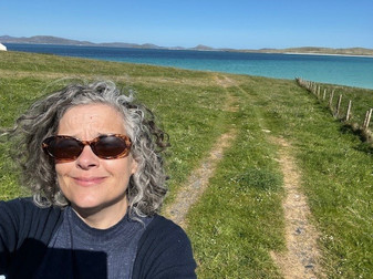 A woman with curly gray hair takes a selfie outdoors on a grassy path leading to a body of water, with clear blue skies in the background.