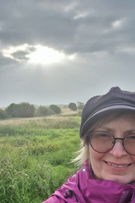 A woman in a purple jacket and dark hat stands in a grassy field with bushes and trees, under a cloudy sky with sunlight streaming through.