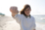 a young girl holding a mole crab in her hand. She is standing on the beach with beach chairs and gentle surf in the background.