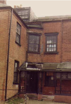 Rear elevation of a brick building showing a corner of that building A sloping roof overhangs downstairs window