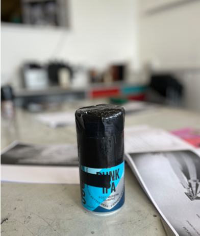 Homemade pinhole camera made from a beer can wrapped in black tape, placed on a table with papers in the background.