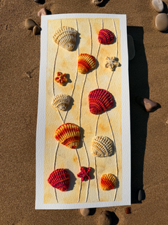 Sandy beach with a small number of stones with a vertical rectangular piece of paper with an embroidered set of sea shells and small stones