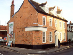 Colour photograph of a brick building with9 windows, three attic windows and a door. Behind it are various building signs/traffic cones