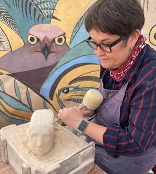A woman sits in front of a busy wall and carves a stone head
