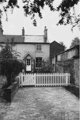 Front of an old property with a roofed front door and a white wooden fence across the front and an empty driveway